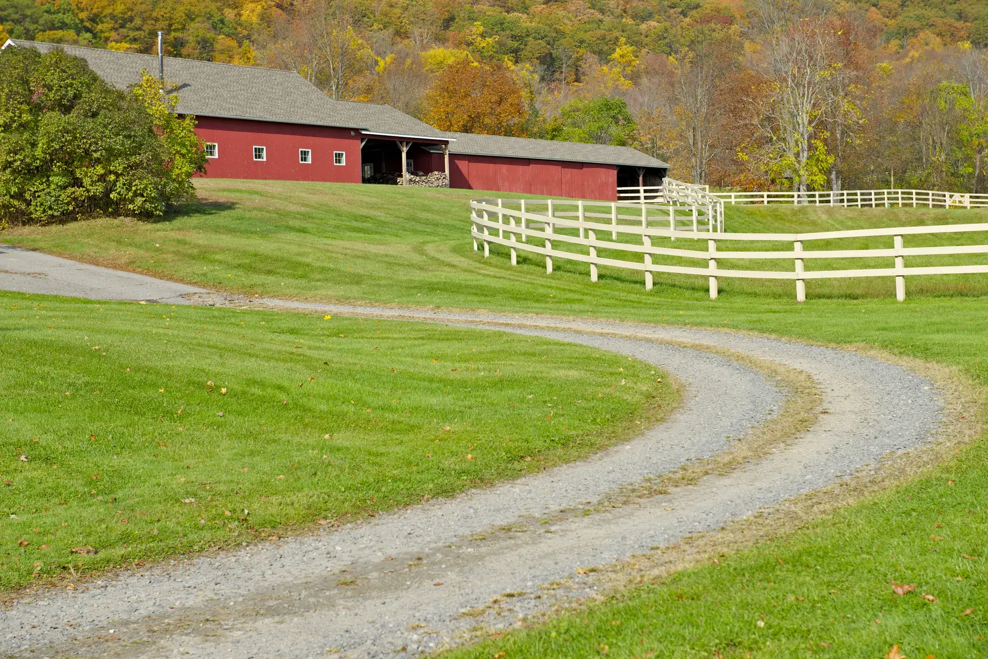 Gravel driveway curving across a mowed lawn past a red barn and white fence