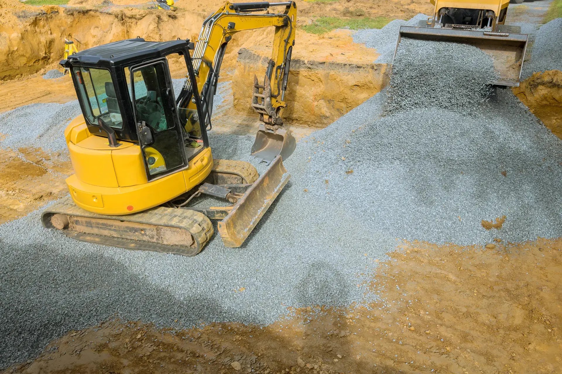 Mini excavator spreading crushed stone with its blade while a loader dumps more behind it