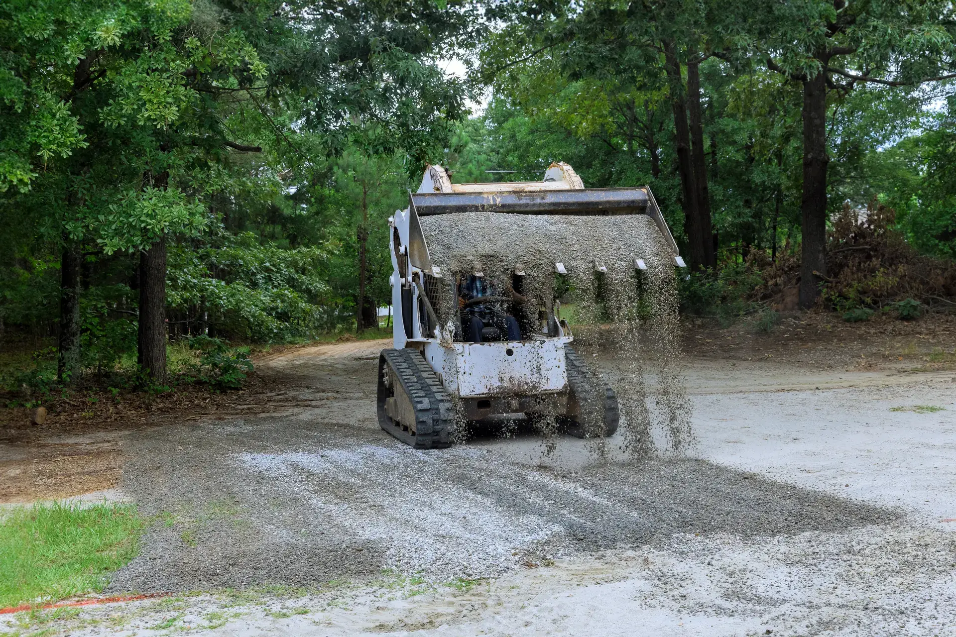 Track loader dumping gravel from its bucket onto a driveway in the woods